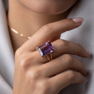 May include: A gold ring featuring a large, rectangular, faceted purple amethyst gemstone set in a four-prong setting. The band of the ring is thin and features a split design where the metal separates into two distinct lines near the stone.