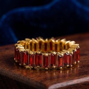 Könnte beinhalten: Ein goldener Eternity-Ring mit einer durchgehenden Reihe tiefroter, im Baguetteschliff gehaltener Edelsteine, die vertikal angeordnet sind. Der Ring besteht aus einer gelbgoldenen Fassung, die jeden einzelnen rechteckigen Stein sicher hält.
