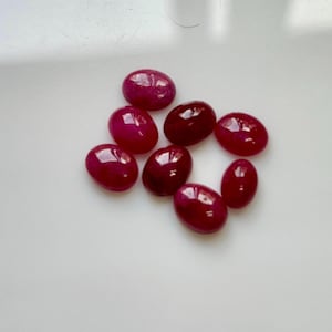 Puede incluir: Colección de nueve gemas ovaladas pulidas en varios tonos de rojo. Las gemas tienen una superficie lisa y brillante, que refleja la luz. Las piedras están dispuestas sobre una superficie blanca, mostrando su color y forma.