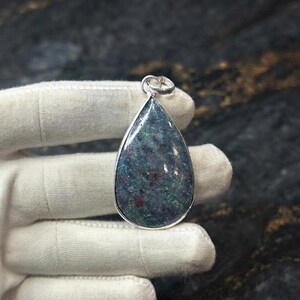 May include: A teardrop-shaped pendant featuring a polished, multi-coloured stone with deep green, blue, and dark red speckled inclusions. The stone is encased in a smooth, polished silver-toned metal bezel setting with a small circular bail at the top.