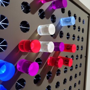 May include: A light brown pegboard panel featuring a grid of circular holes, with several translucent cylindrical pegs in vibrant shades of pink, red, blue, purple, and clear inserted into the openings, creating a colorful and tactile display.