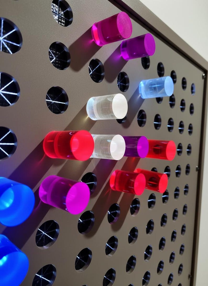 May include: A light brown pegboard panel featuring a grid of circular holes, with several translucent cylindrical pegs in vibrant shades of pink, red, blue, purple, and clear inserted into the openings, creating a colorful and tactile display.