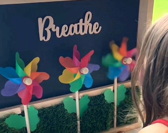 May include: A wooden frame with a chalkboard background that says "Breathe". There are three colorful pinwheels in front of the chalkboard, each with a green stem and leaves. The pinwheels are in a row, and the one on the right is slightly blurred. The frame is sitting on a table with a child's hand reaching towards the pinwheels.