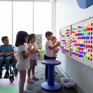 May include: A wall-mounted light board featuring a grid of circular holes filled with translucent, multicolored plastic pegs that glow in shades of red, blue, green, and yellow. The wooden-framed sensory board is mounted on a white wall.