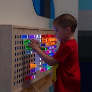 May include: A light-colored wooden sensory wall panel featuring a grid of circular holes. Translucent, colorful cylindrical pegs in shades of red, orange, green, blue, and purple are inserted into the holes, with some pegs illuminated from within.