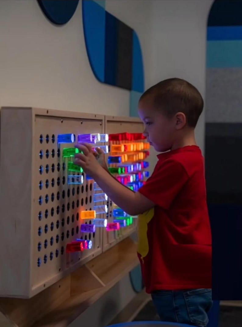 May include: A light-colored wooden sensory wall panel featuring a grid of circular holes. Translucent, colorful cylindrical pegs in shades of red, orange, green, blue, and purple are inserted into the holes, with some pegs illuminated from within.
