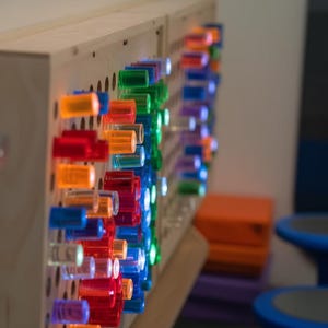 May include: A light-colored wooden sensory wall panel featuring a grid of numerous translucent, cylindrical pegs in vibrant shades of red, orange, blue, and green. The pegs are arranged in rows and columns, creating a colorful, tactile display.