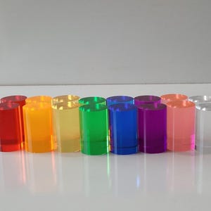 Op de afbeelding: Een set van 12 kleurrijke acrylcilinders in verschillende tinten rood, oranje, geel, groen, blauw, paars, roze en helder. De cilinders zijn in een rij van links naar rechts gerangschikt, met de rode cilinder links en de heldere cilinder rechts.