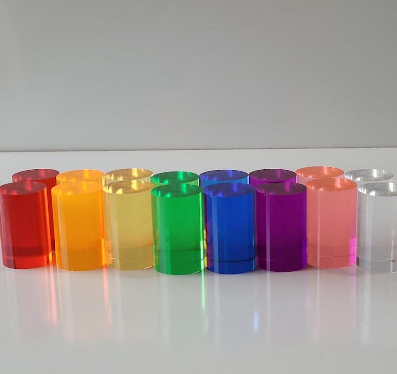 Op de afbeelding: Een set van 12 kleurrijke acrylcilinders in verschillende tinten rood, oranje, geel, groen, blauw, paars, roze en helder. De cilinders zijn in een rij van links naar rechts gerangschikt, met de rode cilinder links en de heldere cilinder rechts.