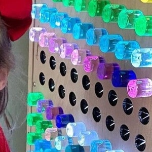 K&ouml;nnte beinhalten: Eine Regenbogen-Sinneswand mit durchsichtigen Plastikr&ouml;hren, die mit farbigem Wasser gef&uuml;llt sind. Die R&ouml;hren sind in Reihen angeordnet, wobei jede Reihe eine andere Farbe hat. Die Wand ist aus Holz und hat einen schwarzen Hintergrund. Eine Kinderhand greift nach einer der R&ouml;hren.