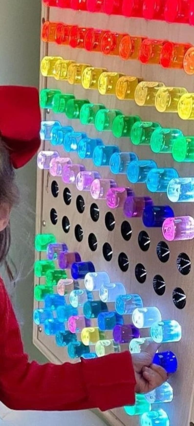 K&ouml;nnte beinhalten: Eine Regenbogen-Sinneswand mit durchsichtigen Plastikr&ouml;hren, die mit farbigem Wasser gef&uuml;llt sind. Die R&ouml;hren sind in Reihen angeordnet, wobei jede Reihe eine andere Farbe hat. Die Wand ist aus Holz und hat einen schwarzen Hintergrund. Eine Kinderhand greift nach einer der R&ouml;hren.