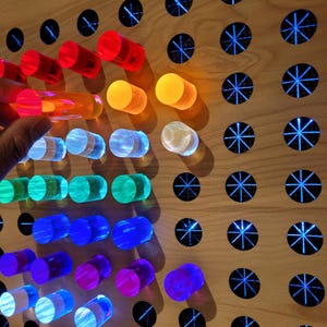 May include: A light-colored wooden board featuring a grid of circular holes, some filled with translucent, colorful cylindrical pegs in red, orange, yellow, green, blue, and purple, while others show a star-shaped pattern illuminated from behind.