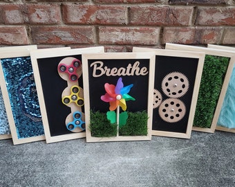 May include: Six wooden framed sensory boards with different textures and activities. The boards include a blue fuzzy texture, blue sequins, three fidget spinners, a green grass texture with a rainbow pinwheel and the word "Breathe", a black chalkboard with gears, green grass texture, and a light blue fuzzy texture.