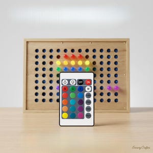 May include: A rectangular light board with a light-colored wooden frame featuring a grid of circular holes. Some holes contain illuminated pegs in red, yellow, blue, green, and purple. A white remote control with twenty-four buttons is in front.