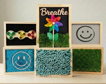 May include: A collection of six rectangular wooden sensory frames. One features the word Breathe with a rainbow pinwheel and artificial grass. Others include three colorful fidget spinners, a smiley face drawing, blue sequins, and green turf.