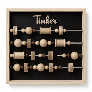 May include: Wooden abacus toy with the word "Tinker" at the top. The abacus has a black background and a light wood frame. The beads are various shapes and shades of brown and tan.