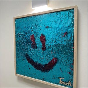 Puede incluir: Una obra de arte mural cuadrada con marco de madera. La obra presenta un fondo de lentejuelas turquesas con un diseño de cara sonriente roja. La palabra "Touch" está escrita en cursiva en la esquina inferior derecha.