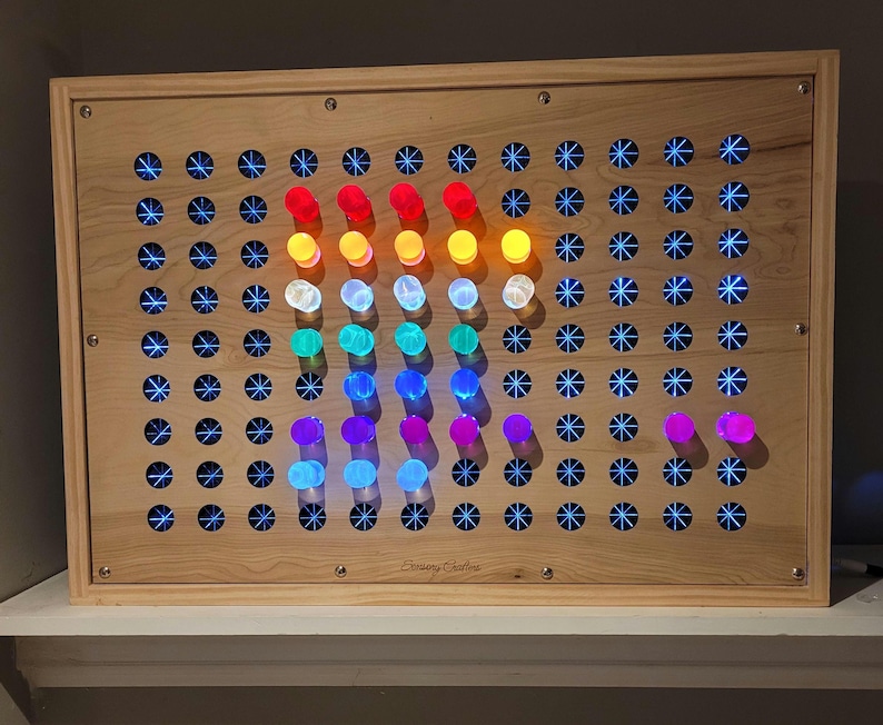 May include: A rectangular light-colored wooden sensory board featuring a grid of circular holes. Many holes contain glowing blue star-shaped lights, while a central section displays a vertical arrangement of colorful pegs in red, orange, yellow, white, green, blue, and purple.