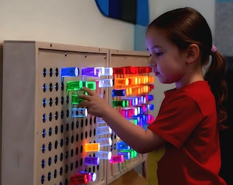 May include: A young person interacts with a light-up sensory wall. The wall features a series of colorful, translucent blocks that emit light in various colors, including red, green, and blue. The blocks are arranged in a grid pattern on a wooden panel.