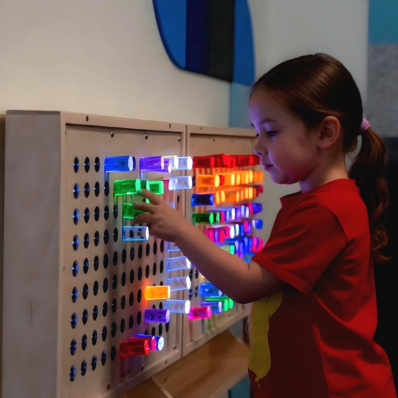May include: A light-colored wooden pegboard wall panel featuring numerous circular holes. Translucent, colorful cylindrical pegs in shades of blue, green, yellow, orange, red, and purple are inserted into the board, emitting a bright, glowing light.