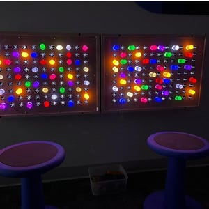 Two Giant Lite-Brite-inspired rectangular wall-mounted light panels featuring a grid of multicolored, glowing cylindrical pegs in shades of red, blue, green, yellow, and white, positioned above two purple stools with round, flat seats.