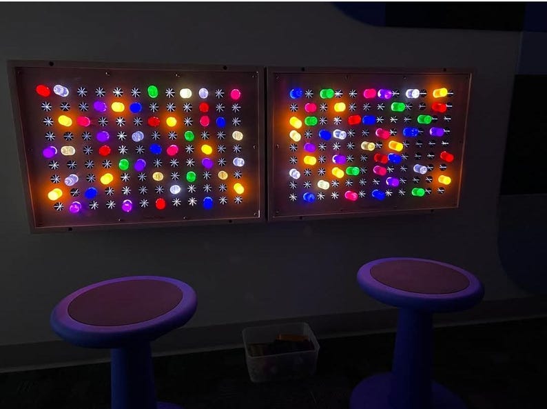 Two Giant Lite-Brite-inspired rectangular wall-mounted light panels featuring a grid of multicolored, glowing cylindrical pegs in shades of red, blue, green, yellow, and white, positioned above two purple stools with round, flat seats.