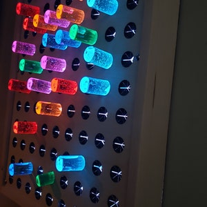 K&ouml;nnte beinhalten: Eine wei&szlig;e, beleuchtete Stecktafel mit bunten, durchsichtigen Stecknadeln. Die Stecknadeln sind in einem Muster angeordnet und werden von hinten beleuchtet, wodurch ein leuchtender Effekt entsteht.