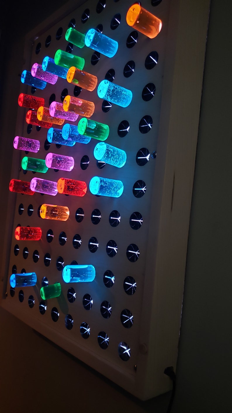 K&ouml;nnte beinhalten: Eine wei&szlig;e, beleuchtete Stecktafel mit bunten, durchsichtigen Stecknadeln. Die Stecknadeln sind in einem Muster angeordnet und werden von hinten beleuchtet, wodurch ein leuchtender Effekt entsteht.