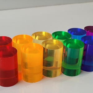 Op de afbeelding: Een set van tien kleurrijke acrylcilinders in een regenboogpatroon. De cilinders zijn helder en doorschijnend, met een lichte glans. De kleuren zijn rood, oranje, geel, groen, blauw, indigo en violet.
