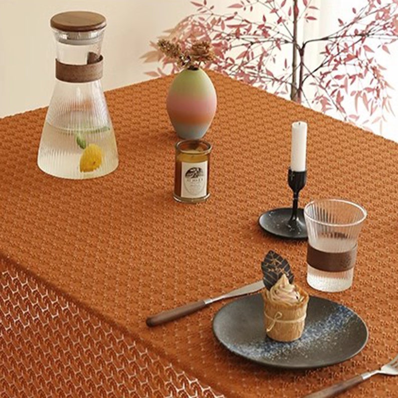以下が含まれることがあります： 茶色の織り模様のテーブルクロス。木製の蓋が付いた透明なガラスのピッチャー、カラフルなデザインの小さな花瓶、ガラスの瓶に入ったキャンドル、黒いキャンドルホルダー、茶色のバンドが付いたグラス、黒いプレートに載せられた小さなデザートがテーブルに置かれている。