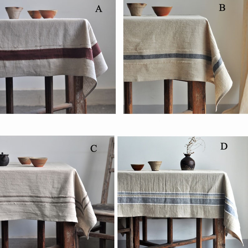May include: Four linen tablecloths with different coloured stripes. The tablecloths are all beige with a single stripe of colour. The stripes are burgundy, blue, blue, and blue. The tablecloths are all laid out on wooden tables.