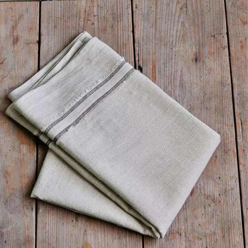 May include: A folded linen tea towel with a black trim. The towel is folded in half and has a subtle texture.