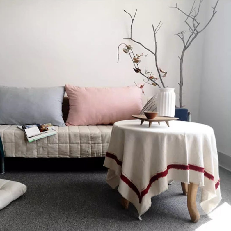 May include: A white linen tablecloth with a red border sits on a wooden table. The table is in front of a sofa with two cushions, one light blue and one pink. A vase with dried branches sits on the table.