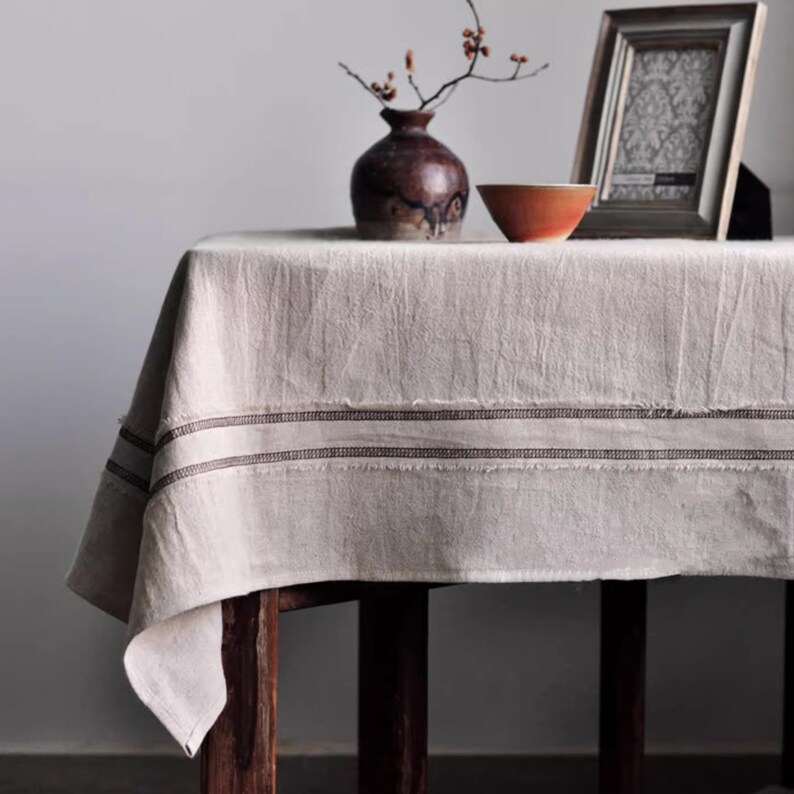 May include: A white linen tablecloth with brown trim, featuring a rustic, farmhouse style. The tablecloth is draped over a wooden table with a vase of dried branches and a small bowl on top.