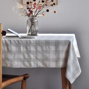 May include: A light gray and white checkered tablecloth draped over a wooden table. A clear glass vase with dried flowers and a book are on the table. The scene is lit by natural light.