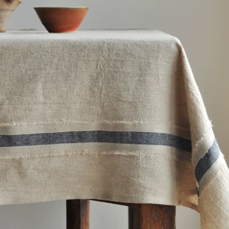 May include: A natural linen tablecloth with a frayed edge and a dark blue stripe running along the bottom. The tablecloth is draped over a wooden table.