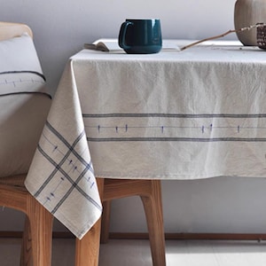 Puede incluir: Un mantel color crema con un diseño de rayas azules y un cojín a juego. Una taza de color azul oscuro está sobre la mesa. El mantel está sobre una mesa de madera, creando un ambiente acogedor y acogedor.