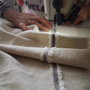 May include: A close-up of a sewing machine stitching a piece of white fabric with a grey stripe. The fabric is folded over and the machine is sewing a straight seam.