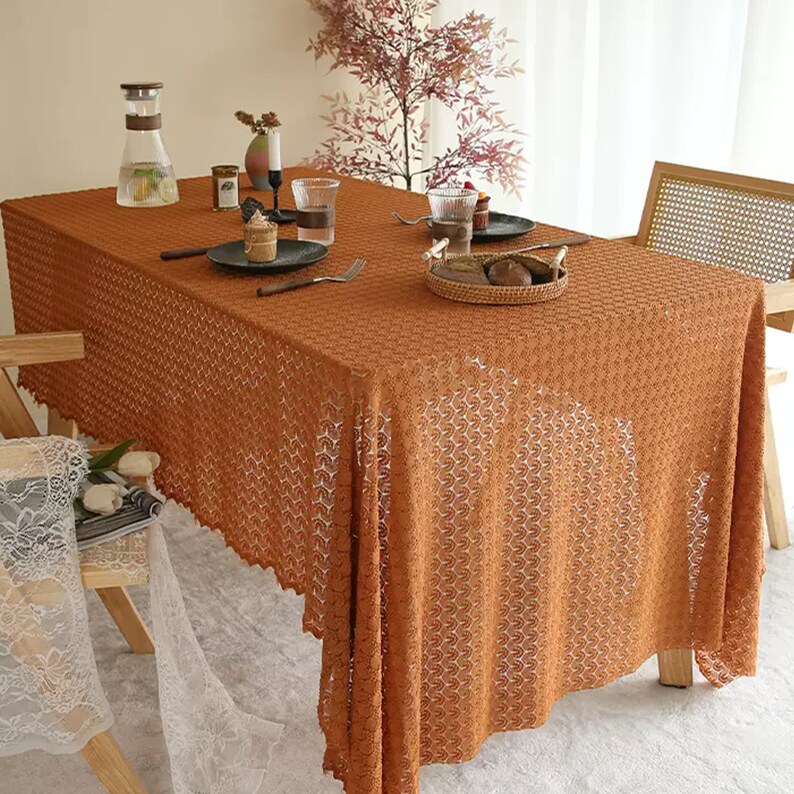 以下が含まれることがあります： 繊細な模様の入った茶色のクロシェのテーブルクロスが木製のテーブルを覆っています。テーブルクロスは波状の縁取りがされており、ピッチャー、グラス、そしてペストリーの載った小さなプレートが置かれています。
