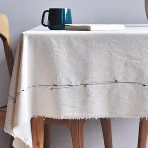 May include: A cream-colored tablecloth with a frayed edge and a black stitched design. A dark teal mug and a small book rest on the table. The tablecloth is draped over a wooden table.