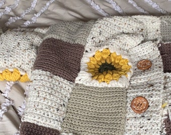 Pode incluir: Um casaco de crochê com um design de patchwork que apresenta quadrados marrons, bege e brancos. Cada quadrado tem um girassol de crochê no centro. O casaco tem botões de madeira.