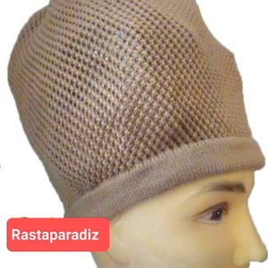 Peut inclure: Un bonnet en tricot beige avec un motif en maille. Le bonnet a un bord roulé et est porté sur une tête de mannequin. Le mot "Rastaparadiz" est imprimé sur un rectangle rouge dans le coin inférieur gauche.