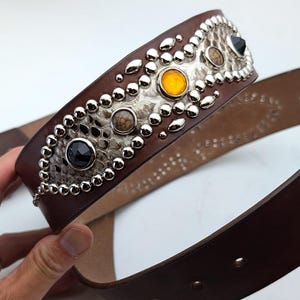 Op de afbeelding: Een bruine leren riem met een decoratief ontwerp van zilver en wit slangenleer. De riem heeft een rij zilveren studs en gekleurde edelstenen.