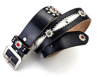 May include: A black leather belt featuring a silver-toned metal buckle, decorative silver studs arranged in geometric patterns, and embedded red circular stones. The belt strap is smooth and dark, with a natural tan underside visible.