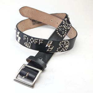Op de afbeelding: Een zwarte leren riem met zilveren studs en een gesp. De riem is versierd met verschillende geometrische vormen en ontwerpen.