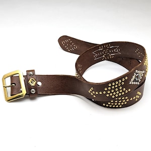 以下が含まれることがあります： ゴールドとシルバーのスタッズと幾何学模様が施されたブラウンの革ベルト。ベルトには真鍮製のバックルが付いています。