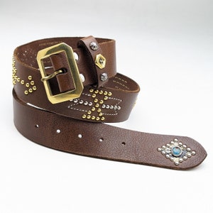 以下が含まれることがあります： 真鍮のバックルと銀のスタッズが付いた茶色の革ベルト。ベルトは幾何学模様で、ターコイズストーンで飾られています。