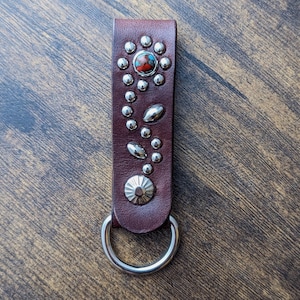 May include: A brown leather key chain with a silver flower design and a silver ring. The flower has a red centre and silver petals.