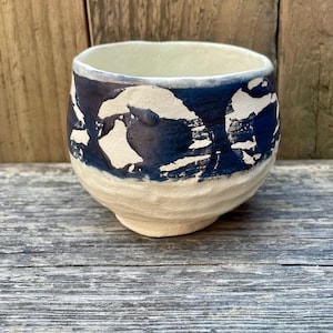 Peut inclure: Tasse en céramique faite à la main, avec une base crème et un motif abstrait bleu foncé autour du bord. La tasse a une forme arrondie et une surface légèrement texturée, mettant en valeur le savoir-faire artisanal. Une pièce unique.