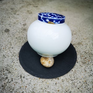 Può includere: Un vaso di ceramica bianca con un coperchio floreale blu e bianco, appoggiato su un disco di ardesia nero. Il vaso poggia su un piccolo sasso rotondo di colore marrone chiaro.